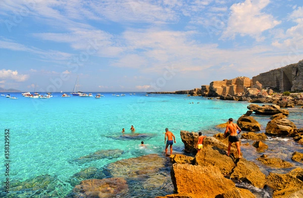 Fototapeta paradise clear torquoise blue water with boats and cloudy blue sky in background in Favignana island, Cala Rossa Beach, Sicily South Italy.