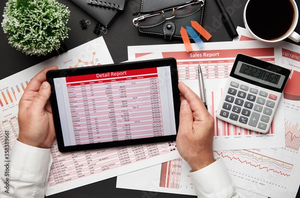 Fototapeta Top view of businessman's hands working with tablet pc and financial reports. Modern black office desk with notebook, pencil and a lot of things. Flat lay table layout.
