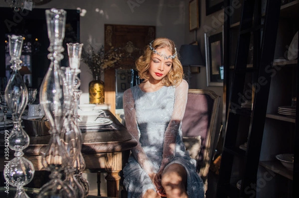 Obraz Beautiful blond girl with vintage make up and hairstyle sitting near table with sunny rays on face and jewelry decoration on head
