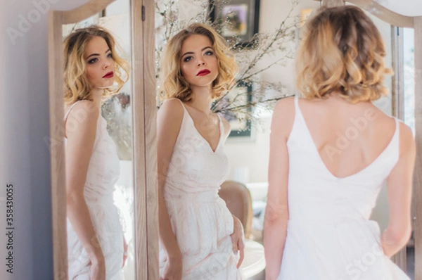 Obraz Beautiful blond girl with vintage make up and hairstyle staying near triple mirror with sunny rays on face