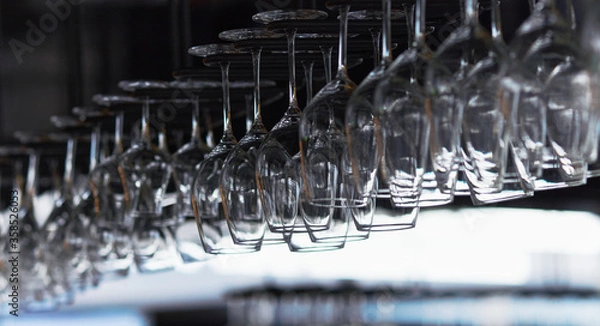 Fototapeta Many empty wine glasses hanging upside down in a row on a special rack over the counter of a bar.