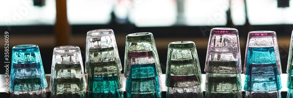 Fototapeta Many colorful backlit bar glasses stacked in a row on the counter of a restaurant.