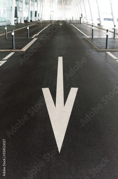 Fototapeta White arrow street sign on dark asphalt, pointing downwards at a parking zone with futuristic white architecture columns in the blurry background.