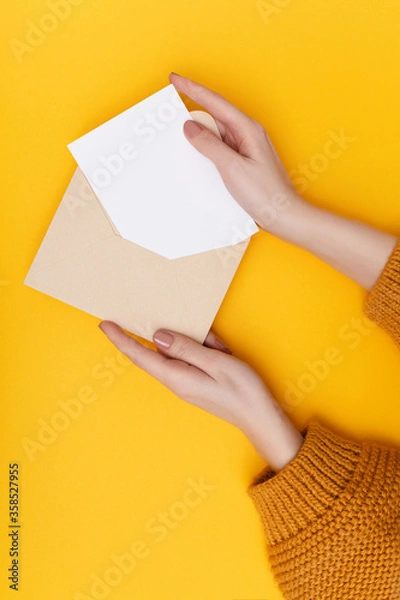 Fototapeta Biege envelope with blank white postcard in woman's hands on the yellow background. Mail concept