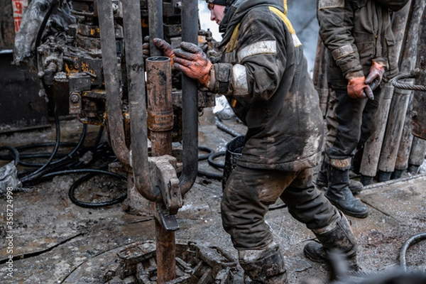 Fototapeta Two working drillers in a uniform, in a helmet and goggles, install drill pipes after lifting them from an oil well after drilling. The concept of a working person.