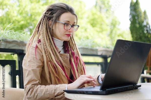 Fototapeta Concentrated female journalist in eyeglasses for vision correction and long dreadlocks hair is writing a questions for next interview using laptop computer while sitting in coffee shop outdoors.