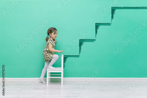 Fototapeta The child climbs onto a hand-drawn ladder. The beginning of a career concept.Funny little girl on a turquoise background with a place for text.