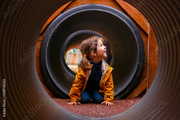 Fototapeta A little girl climbs in the pipes. Modern and safe children's Playground. Round and long tunnel. Overcome obstacles. Children's curiosity and discovery of everything new.