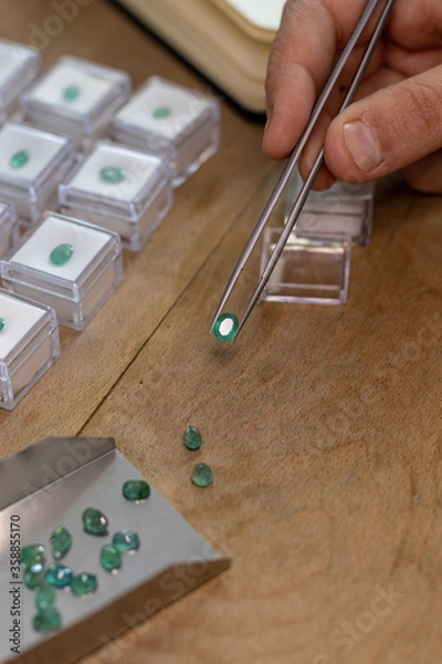 Obraz Hand holding a emerald gemstone with jewelry forceps. Gemologist workspace.