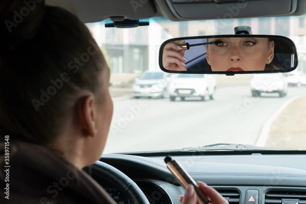 Fototapeta Beautiful female driver makes-up in the car and putting mascara on her eyes. Young lady looking at rearview mirror in the car. Focus on the mirror