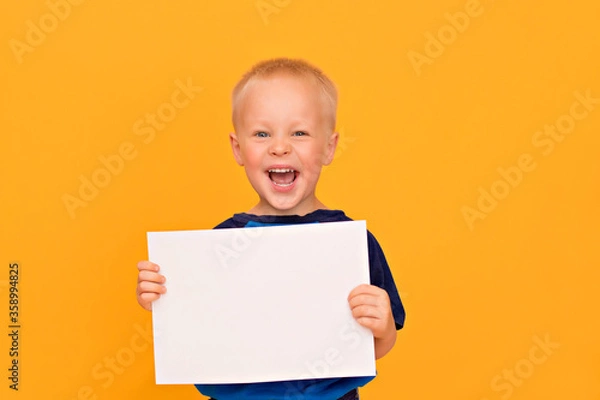 Obraz Happy boy with a smile holds a blank white sheet. Yellow background copy space
