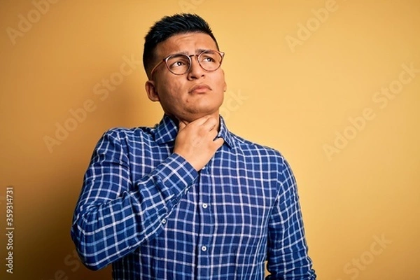 Fototapeta Young handsome latin man wearing casual shirt and glasses over yellow background Touching painful neck, sore throat for flu, clod and infection