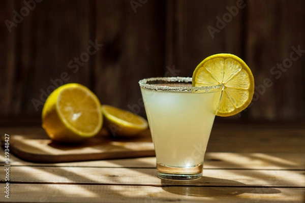 Fototapeta summer alcoholic yellow frosted drink in a transparent shaped glass with sugar on the rim and a round ripe juicy slice of lemon on a wooden table and round lemons in the background