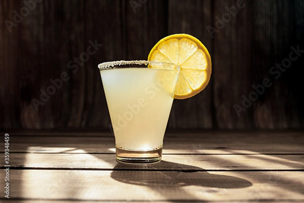 Fototapeta summer alcoholic yellow frosted drink in a transparent shaped glass with sugar on the rim and a round ripe juicy slice of lemon on a wooden table, photo for the bar menu