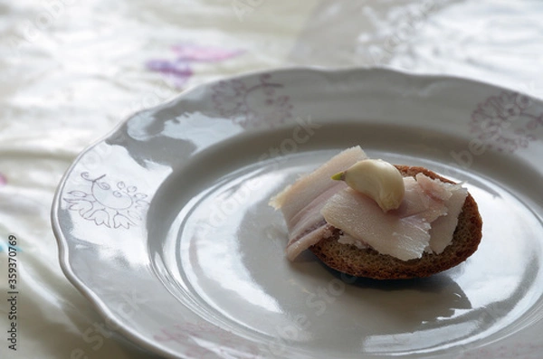 Fototapeta Slices of lard and a clove of garlic on a small hump of gray bread. Snack on a plate closeup.