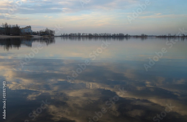 Fototapeta Landscape in the bay at the Dnieper river, Ukraine. Reflection of blue sky and light pink clouds in calm water. Buildings and trees on the shore on the horizon. Quiet March spring evening.