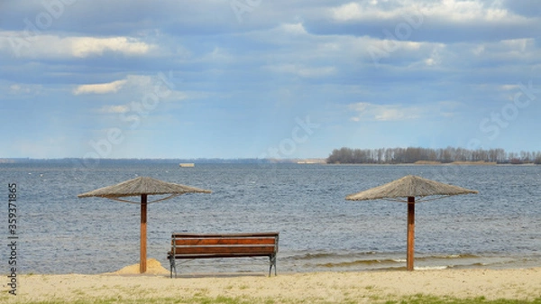 Fototapeta An empty bench and two reed umbrellas on the sandy shore of a pond against water in the spring in March. Blue cloudy sky. Forest on the horizon.