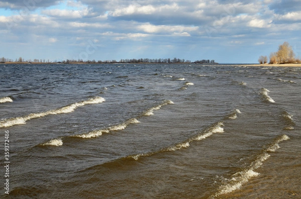 Fototapeta A series of running waves with white crests in shallow water in the foreground. Cloudy sky over the horizon. Landscape on the right bank of the Dnieper River in March, Ukraine.