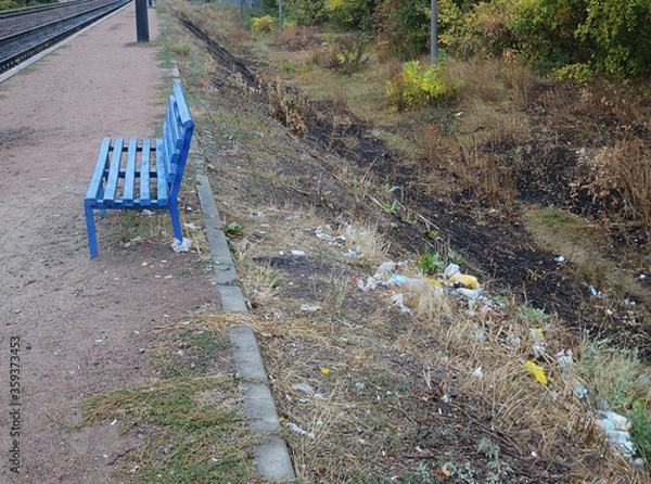 Fototapeta Bench at the polluted platform of a suburban railway station. Trash on the slope of the embankment near the bench.