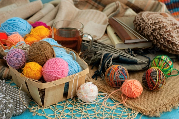 Obraz Needlework in the form of knitting. Knitting yarn, knitted items, a warm shawl and tea create coziness. Colorful multi-colored balls of yarn in a basket and on a table.