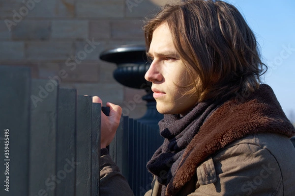 Fototapeta Tense young handsome guy, gripping his hand  onto an iron fence and looking serious into the distance on a sunny day in winter.