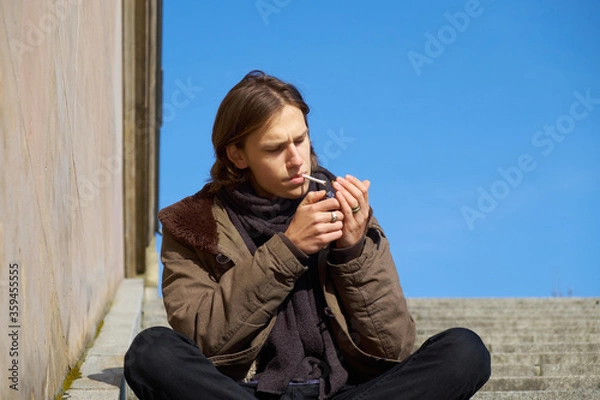 Fototapeta Young casual handsome guy with brown hair lighting up a self rolled cigarette on a sunny winters day, while sitting on an ancient staircase in Berlin city.