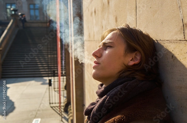 Fototapeta Young casual handsome guy with brown hair exhaling the white smoke of a cigarette on a sunny winters day, while leaning on a wall in Berlin city.