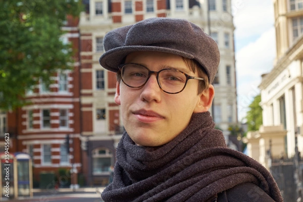 Fototapeta Young casual handsome guy wearing brown glasses and a trendy gray ivy flat cap, glancing amused to the camera in an urban setting in London city.