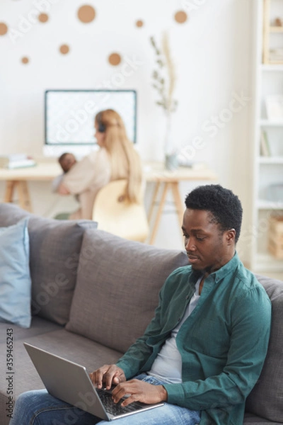Fototapeta Vertical portrait of modern interracial family working from home, focus on African-American man using laptop in foreground, copy space