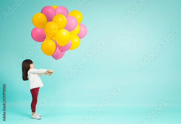 Fototapeta Little girl holding helium balloons on plain background