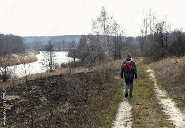 Fototapeta A girl in an autumn jacket, hat and with a backpack takes a walk on nature in early spring. He looks at the vast expanses and enjoys the view.