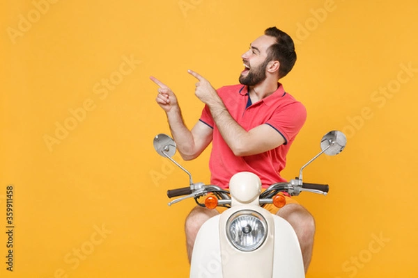 Fototapeta Cheerful young bearded man guy in casual summer clothes driving moped isolated on yellow background studio. Driving motorbike transportation concept. Mock up copy space. Point index fingers aside up.