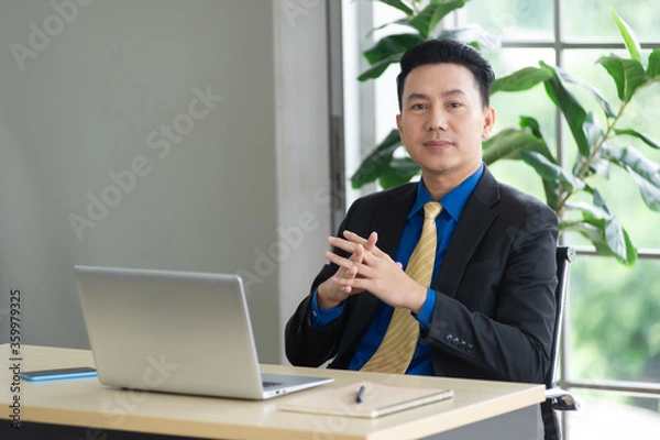 Obraz Portrait of middle age Asian businessman wearing formal black suit sitting on chair, there are laptop and smartphone at his desk, looking at camera, working in workplace