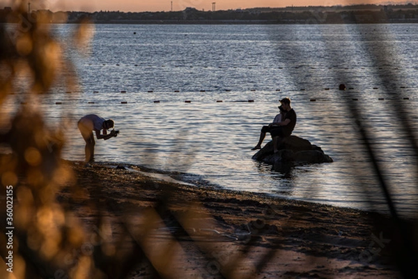 Obraz In the distance, a photographer takes pictures of young people on the shore of a sandy beach, calm sea at sunset in the resort town of Gelendzhik. Russia, black sea coast