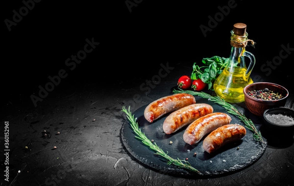 Fototapeta Grilled sausage with the addition of herbs and vegetables on a dark background