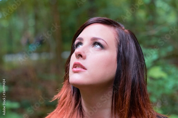 Fototapeta Thoughtful young woman with gray eyes looking up to the sky, wearing nose jewellery, with a stunning hair style in color tones of crimson red and orange, standing in a forest.