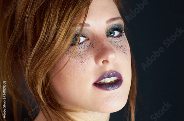 Fototapeta Close up of a young red headed woman with dark make up mascara, purple lip color and metallic colorful glitter under her gray eyes with a charming glance.