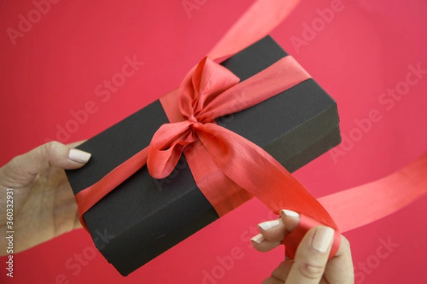 Fototapeta Girl holds a gift box and unties the bow. a girl opens a gift box. Gift box on a red background. Girl unties the tape from the box.
