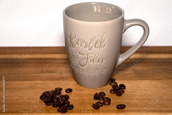 Fototapeta Coffee cup says "Love and Fika" on a wooden table with coffebeans infront on a white background