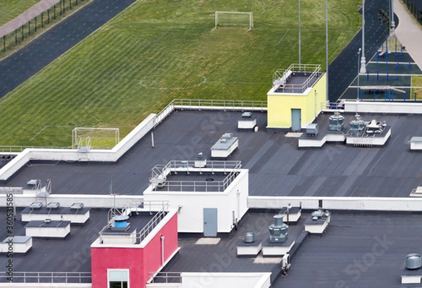 Fototapeta Drone point of view dark flat roof with air conditioners and hydro insulation membranes on top of a modern apartment building with green sport field residential area.