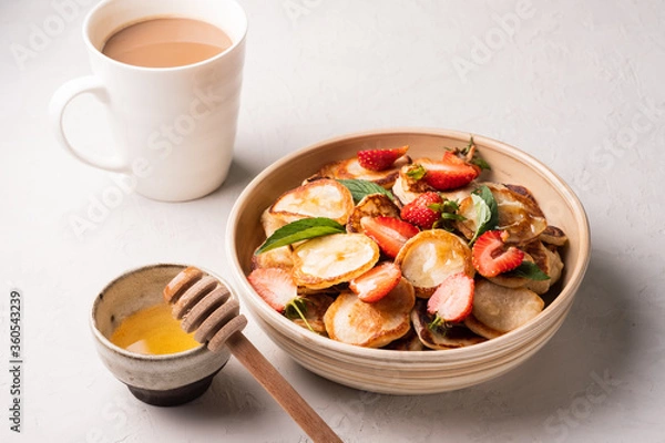 Fototapeta Bowl with tiny cereal pancakes with strawberries, mint and honey on a gray concrete background. Fashionable food. Mini cereal pancakes. Pancake porridge.