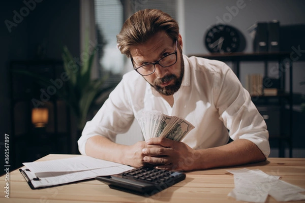 Fototapeta Confident businessman in glasses and formal outfit holding money cash while sitting at table with documents and calculator. Mature man looking at camera while working with financial report.