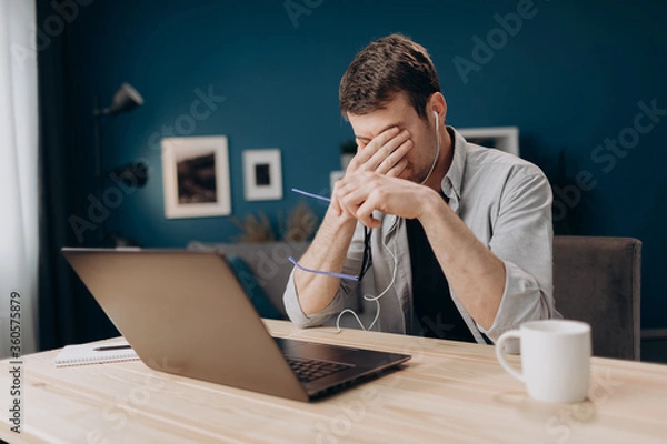 Fototapeta Young man in casual outfit rubbing his eyes because of long working day with portable computer. Bearded freelancer feeling tired while using laptop at home.