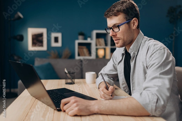 Fototapeta Side view of bearded man in eyeglasses and casual shirt making notes while working on laptop at home. Young guy sitting at table and using modern device.