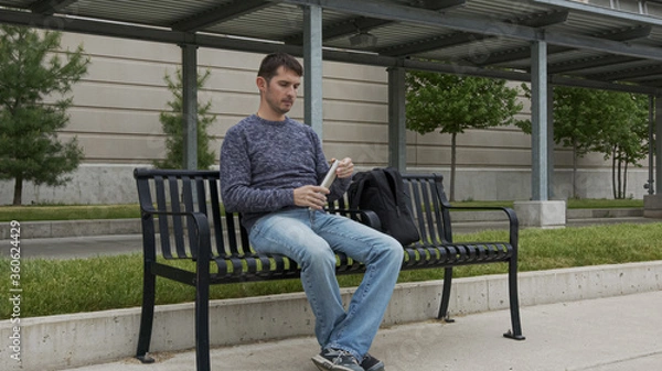 Fototapeta High quality picture of a young man who is sitting on a metal bench outside of train and bus station and drinking water from the stainless steel thermos which he took from the backpack.