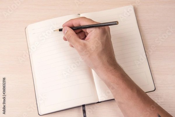Fototapeta open notebook with black pencil in hand on wooden background top view, school notepad and male hand on a table, a desktop in the office