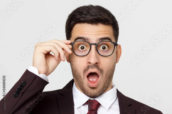 Fototapeta Young businessman looking at camera with surprise expression, shouting WOW with open mouth, isolated on gray background
