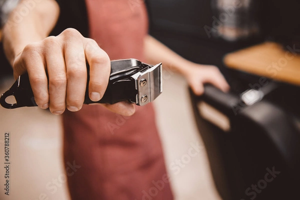 Fototapeta Barber holds hair clipper in modern barbershop for men, brown retro