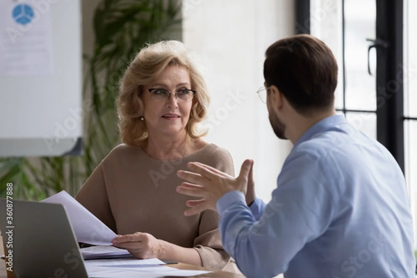 Fototapeta Mature businesswoman wearing glasses discussing project strategy with business partner at meeting, holding financial documents, colleagues working together, sitting at table in boardroom