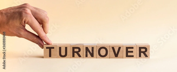 Fototapeta The hand puts a wooden cube with the letter T from the word TURNOVER. The word is written on wooden cubes standing on the yellow surface of the table. Concept.